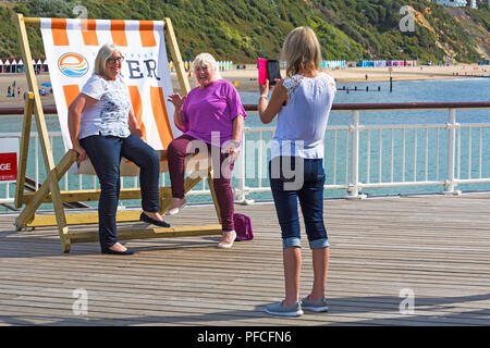 Bournemouth, Dorset, UK. 22nd Aug, 2018. UK weather: warm and sunny at Bournemouth beaches as sunseekers head to the seaside to enjoy the weather. Friends pose for a photo in the big deckchair on Bournemouth Pier. Credit: Carolyn Jenkins/Alamy Live News Stock Photo