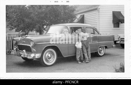 Black and white photograph, showing two small, smiling, blonde boys, likely brothers, standing in front of the driver's side door of a two-tone, hardtop Chevrolet Bel Air, that is parked in a driveway, with a small, light-colored house, tree, and picket fence visible in the background, likely photographed in Ohio, August, 1955. () Stock Photo