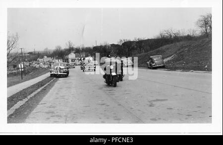 Black and white photograph, showing the beginning of a parade or motorcade, possibly celebrating the end of World War II, headed by a pair of police motorcycles, with several vintage cars (one festooned) in the background, and leafless trees visible on either side of the road, likely photographed in Ohio, 1945. () Stock Photo