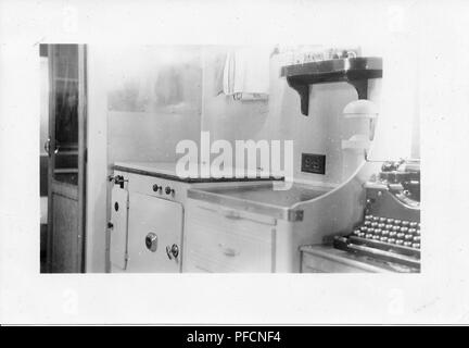 Black and white photograph, showing the cramped interior of a Mid-century caravan or trailer home, with part of a black typewriter visible next to a small, tiled kitchen area with a vintage oven, likely photographed in Ohio in the decade following World War II, 1950. ()