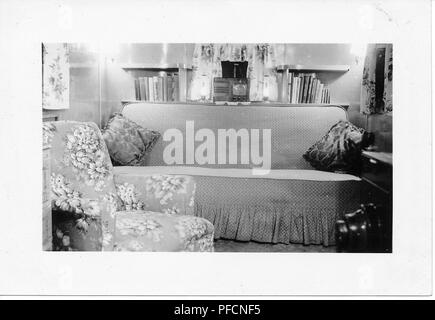 Black and white photograph, showing the cramped interior of a vintage caravan or trailer home, dominated by a floral easy chair, and a chintz-covered sofa, with a small radio, and books on shelves visible in the background, likely photographed in Ohio in the decade following World War II, 1950. ()