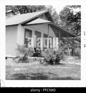 Black and white photograph, showing the front of a small house, with bushes screening several chairs on a front porch, and foliage visible in the background, photographed in Ohio, June, 1956. () Stock Photo