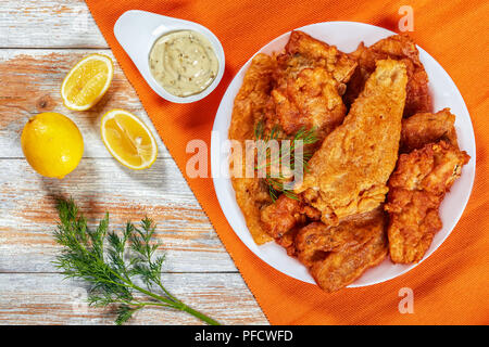 delicious crispy pan fried Beer Battered sea fish fillets on white platter on old wooden table. tartar sauce in gravy boat, dill and lemon on backgrou Stock Photo