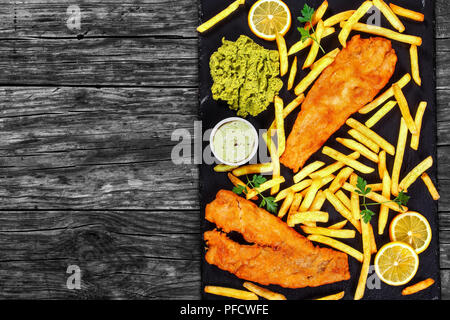 delicious crispy fish and chips on black slate plate with tartar sauce, mushy pea, lemon slices on dark wooden background, view from above, blank spac Stock Photo
