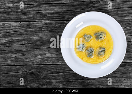 hot yummy  creamy polenta with chunks of melted gorgonzola cheese on white wide rim dish on old dark wooden table, view from above Stock Photo