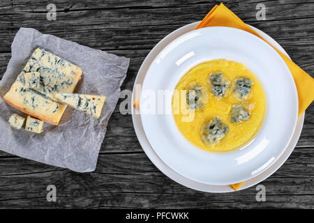 hot delicious creamy thick polenta with chunks of melted gorgonzola cheese on plate with napkin. piece of gorgonzola on paper on old dark wooden table Stock Photo