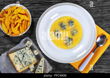 hot delicious creamy polenta with melted gorgonzola cheese on plate with napkin and spoon. piece of gorgonzola on paper, saucer with cornmeal  spicy b Stock Photo
