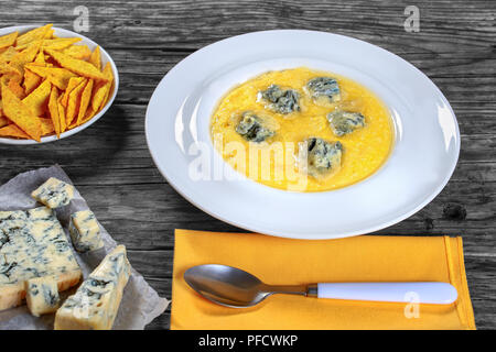 hot delicious creamy polenta with melted gorgonzola cheese on white plate on dark table with spoon on napkin. piece of gorgonzola on paper, biscuits i Stock Photo