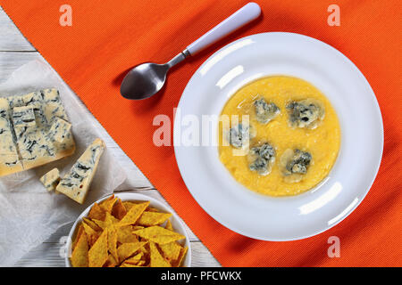 hot delicious creamy polenta with melted gorgonzola cheese on white plate on orange table mat with spoon. piece of gorgonzola on paper, biscuits in bo Stock Photo