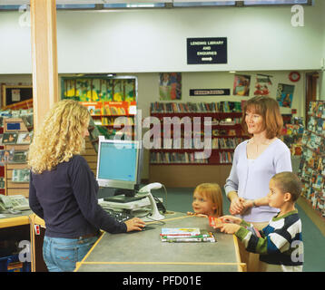 Woman, boy and girl smiling at librarian standing behind library reception desk with hand on computer mouse, side view. Stock Photo