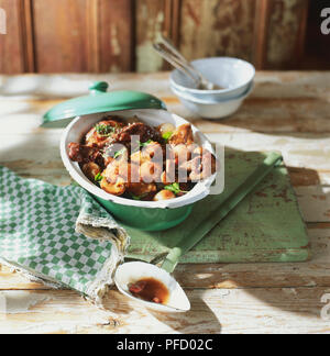 Casserole dish, lid resting on rim, with pieces of chicken, button onions, mushrooms and parsley leaves, sauceboat with juices in front, stacked bowls and spoons in background, front view. Stock Photo