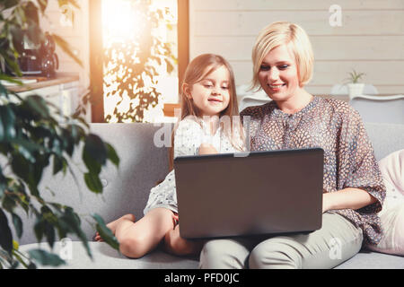 Pretty blonde mother and cute little brunette girl sitting on the sofa and using the laptop Stock Photo