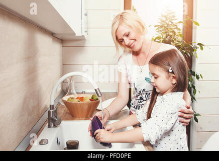 Beautiful blond mother and cute brunette little daughter wash dishes on the light kitchen Stock Photo