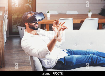 Multicultural bearded man laying on the sofa and using virtual reality goggles in the modern flat Stock Photo