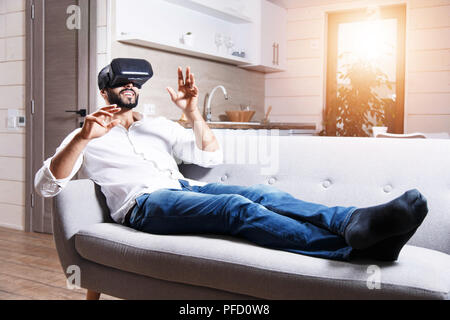 Multicultural bearded man laying on the sofa and using virtual reality goggles in the modern flat Stock Photo