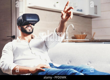 Multicultural bearded man laying on the sofa and using virtual reality goggles in the modern studio kitchen Stock Photo