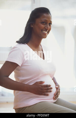 Woman with dark skin and straight, long black hair, in middle to later stages of pregnancy, sitting on window sill and stroking belly with both hands, smiling, side view Stock Photo