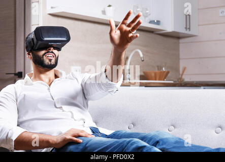 Multicultural bearded man laying on the sofa and using virtual reality goggles in the modern flat Stock Photo