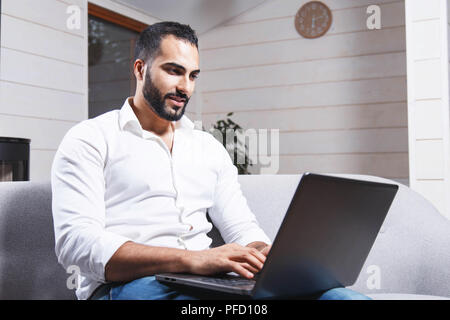 Handsome young enterpreneur, bearded man wears white shirt sitting on the sofa and working with laptop in the modern flat Stock Photo
