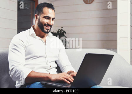 Handsome young businessman, bearded man wears white shirt sitting on the sofa and working with laptop in the modern flat. Stock Photo