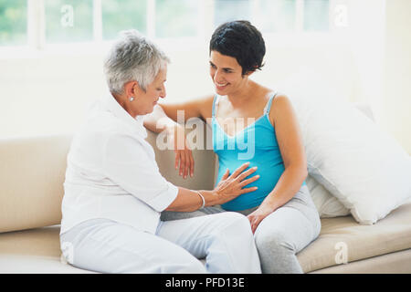 Middle-aged woman touching the belly of smiling pregnant woman, side view. Stock Photo