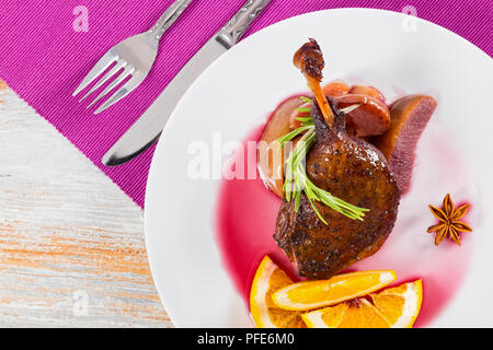 fried duck legs baked in oven in red wine with rosemary,orange and apple slices on dish, on table mat,  on rustic table, french cuisine, close-up, vie Stock Photo
