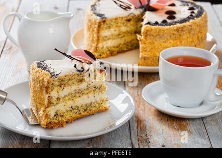sponge cake with poppy seeds layered with cream with a cut out piece on cake shovel on white plate close-up with jug of milk, cup with black tea on sa Stock Photo