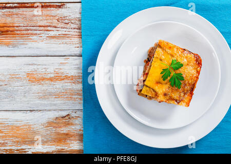 portion of delicious moussaka decorated with parsley cooked for authentic recipe, on plates on table mat, on white peeling paint  boards, view from ab Stock Photo