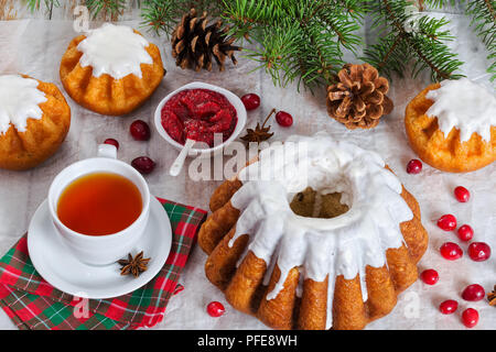 delicious  rum cakes with sugar glaze and cranberry on parchment paper with cup of tea on table napkin and cranberry jam, spruce branches and pinecone Stock Photo