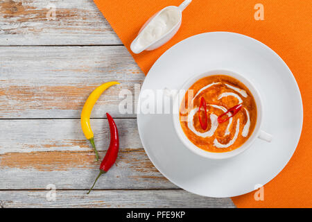 hot colorful Thai Pumpkin Soup with red thai paste, chili sauce and heavy cream in white cup on plate on table mat with cream in gravy boat, yellow an Stock Photo