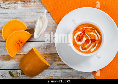 hot colorful Thai Pumpkin Soup puree with red thai paste, chili sauce and heavy cream in white cup on plate on table mat with cream in gravy boat and  Stock Photo