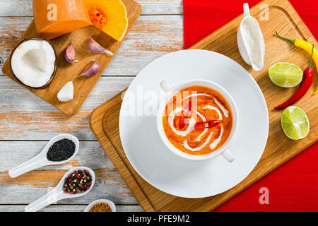 Thai Pumpkin Soup puree decorated with swirl in a little cream and chilli sauce in white cup on plate on chopping board with  ingredients on  backgrou Stock Photo