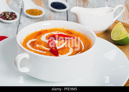 Thai Pumpkin Soup puree decorated with swirl in a little cream and chilli sauce in white cup on plate on chopping board with  lime slice and heavy cre Stock Photo