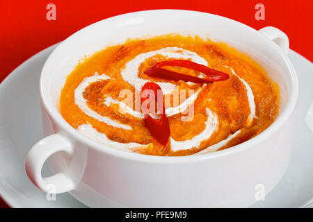Thai Pumpkin Soup puree decorated with swirls in a little cream and red paste, topped with some chopped red chillies. in white cup on orange table mat Stock Photo