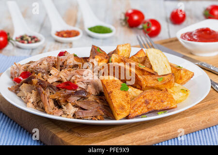 pulled slow-cooked pork shoulder grilled in oven with fried potato wedges on white plate  with tomato sauce on chopping board on wooden table, close-u Stock Photo
