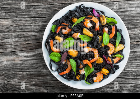 seafood black noodles salad with prawns, mussels, fresh green spinach, lettuce, arugula and mint on white dish on dark wooden table, view from above