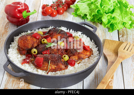 rice with grilled chicken thighs, red bell pepper, green olives and rosemary in cast iron stewpan on white old wooden table, vegetables and lettuce on Stock Photo