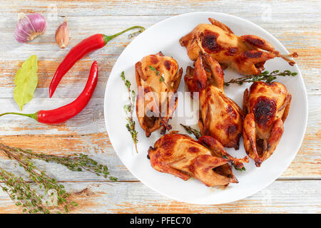 grilled whole quails with thyme on dish on white wooden background with chili peppers, bay leaf and garlic, view from above Stock Photo