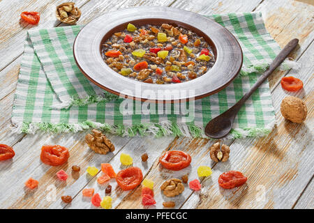 Sweet wheat kutia with nuts, poppy seeds, dried fruits. succade and honey in clay bowl with spoon on kitchen cloth on wooden planks. with ingredients  Stock Photo