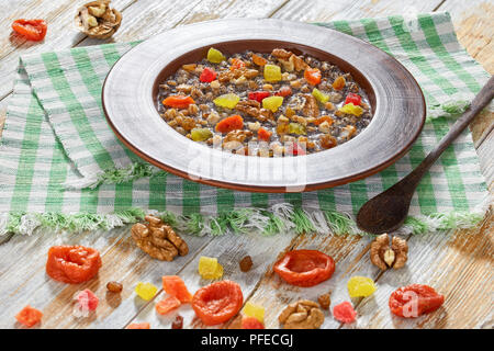 Sweet wheat dessert kutia with nuts, poppy seeds, dried fruits, succade and honey in clay bowl with spoon on table napkin on wooden planks. with ingre Stock Photo