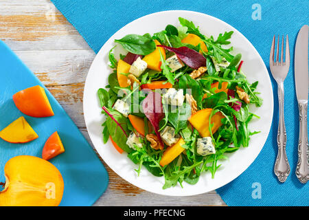 persimmon, blue cheese, spinach, arugula, lettuce leaves and walnuts healthy salad on white platter. slices of fresh sweet persimmon on chopping board Stock Photo