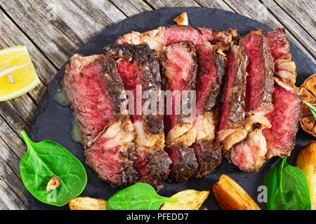 medium-rare grilled rib-eye steak with garlic and rosemary cut in slices on black slate plate with spinach leaves and fried potato, on dark wooden bac Stock Photo