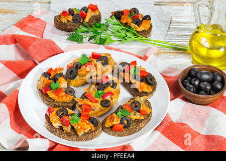 whole grain rye Crostini with pieces of mackerel fish, rings of black olives, red bell pepper on white plate on kitchen cloth, bottle with cooking oil Stock Photo