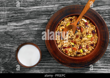 Homemade granola with honey, oatmeal, cashew nut, almond, pistachio, raisin and cranberry in clay bowl with wooden spoon, cup with milk on old dark ta Stock Photo