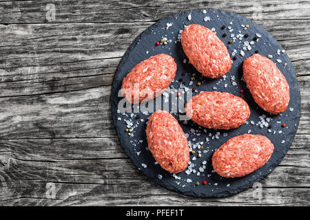 fresh minced raw meat cutlets on slate plate with kosher sea salt and mix of peppers on old dark wooden boards, view from above Stock Photo