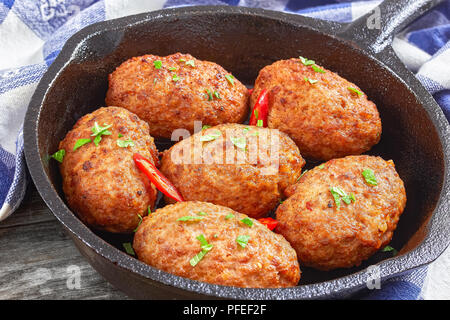 juicy delicious meat cutlets in iron frying pan sprinkled with chili pieces and green herbs. tomato, bay leaves and onion on old rustic table with tow Stock Photo