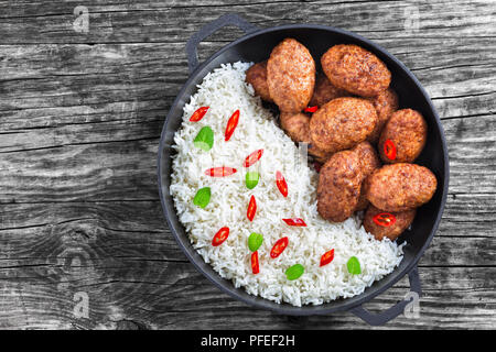 rice and juicy delicious meat cutlets sprinkled with pieces of chili  and green mint in iron stew pan old rustic boards, view from above, close-up Stock Photo