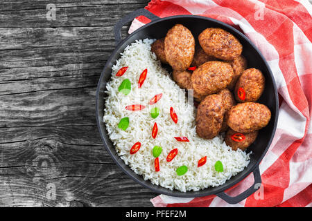 rice and juicy delicious meat cutlets  sprinkled with chili pieces and green herbs in iron stew pan on kitchen towel, view from above Stock Photo