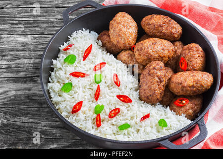 rice and juicy delicious meat cutlets  sprinkled with chili pieces and green herbs in iron stew pan on kitchen towel. chili peppers on old rustic boar Stock Photo