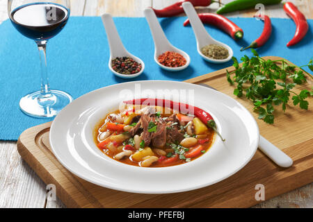 delicious hot beef meat soup with root vegetables, potato, beans, chili and tomato sauce in white plate on chopping board, glass with red wine and spi Stock Photo